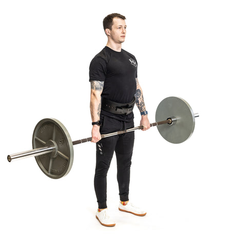 A tattooed person in black clothing performs a deadlift with the Bells of Steel Hydra Seven Ring Bar, gripping its aggressive knurling while standing upright against a white background.