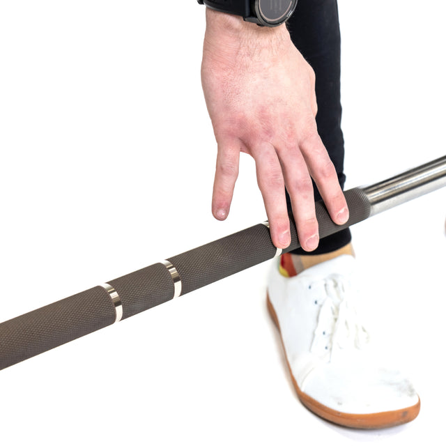 Sporting white sneakers and a smartwatch, a lifter confidently grips the Bells of Steel Hydra Seven Ring Bar with one hand, readying for a powerful lift. The shot emphasizes the exact hand placement on the barbell, set against a clean white backdrop.