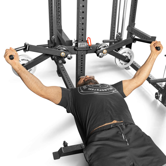 A person uses the Bells of Steel Shoulder Boulder/Chest Fly Attachment, performing a chest fly on a bench for a V-taper look. Gripping the adjustable pulleys' handles, they wear a black shirt and shorts in the gym's space-saving rack setup.