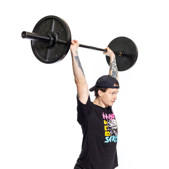 A person, adorned with tattoos on their arms and dressed in a black cap and graphic t-shirt, hoists the Bells of Steel Short Utility Barbell overhead against a plain white backdrop.