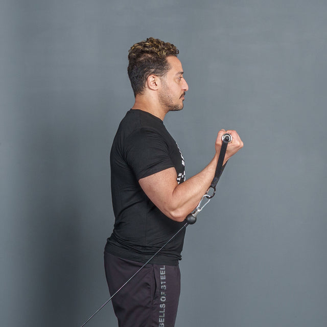 A man in black attire performs a standing cable bicep curl against a gray background, using a Bells of Steel Aluminum Handle with Bearings in one hand and bending his elbow to lift the weight.