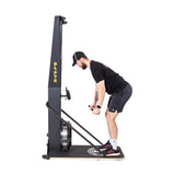 A man in athletic wear uses the Bells of Steel Blitz Ski Trainer, gripping its handles and pulling downward as he stands on the wooden platform, leaning forward with bent knees for a cross-country skiing workout.