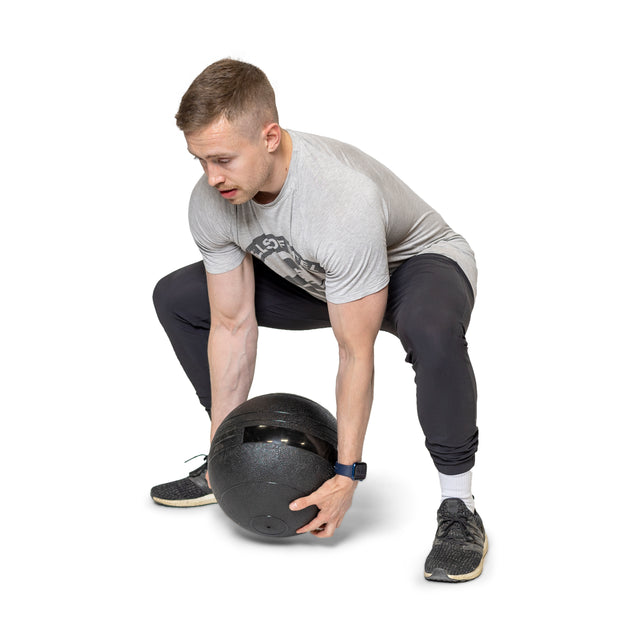 A man in a gray tee and black pants squats while lifting a large black Bells of Steel Slam Ball to build explosive power. He's wearing black sneakers and a smartwatch to track cardio workouts, set against a plain white background.