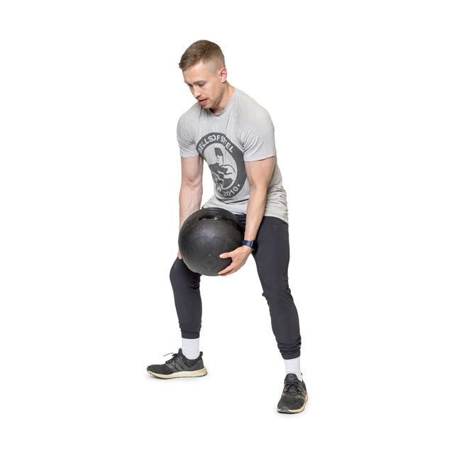 A man in a Bells of Steel gray T-shirt and black pants exercises with a large Slam Ball. With feet apart, he bends forward slightly, holding the ball with both hands, focused on channeling explosive power against a white background.