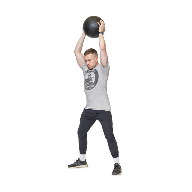 Wearing athletic attire, a person exercises with a Bells of Steel Slam Ball raised overhead, standing with feet apart on a white background. Their focused and determined look showcases the explosive power of their intense workout.