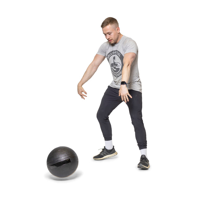 A person in a gray T-shirt, black pants, and sneakers stands ready with arms outstretched towards a Bells of Steel Slam Ball on the floor, indicating they are prepared for explosive cardio workouts. The background is white.