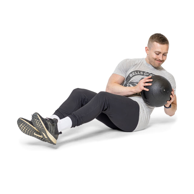 A man performs a powerful Russian twist with feet off the ground, smiling slightly while holding Bells of Steel Slam Balls. He's dressed in a gray t-shirt and black pants against a white background.