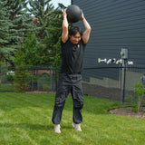 Wearing black clothing, a person stands on the grass of a backyard gym, lifting a medicine ball overhead with both hands as they prepare to slam it down as part of an outdoor workout using the Bells of Steel Ultimate Backyard Training Bundle.