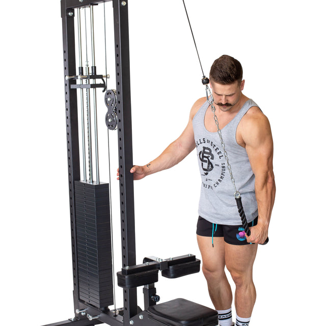 A person performing tricep pushdowns is focusing on their muscles using a cable machine. They are wearing a gray tank top and black shorts, complemented by white socks and sneakers. The workout setup includes a stack of weights and the Bells of Steel Tricep Pulldown Rope – Single for an effective exercise session.