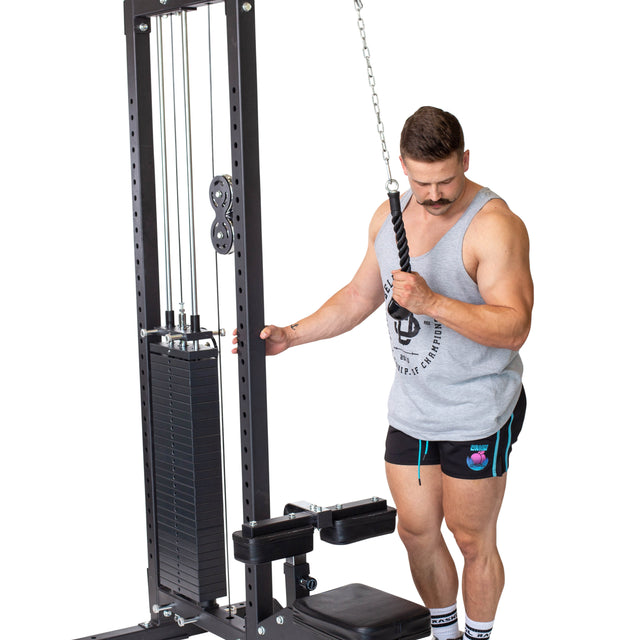 A man in a gray tank top and shorts stands by the cable machine at the gym, gripping the Bells of Steel Tricep Pulldown Rope – Single. He is preparing for a tricep-focused workout, with visible weights stacked on the machine.