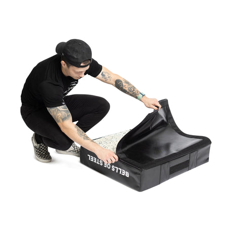 A tattooed individual in a black cap and t-shirt crouches on a speckled floor, lifting the flap of a Bells of Steel Deadlift Pad - Replacement Cover made from black 600 Denier Polyester.