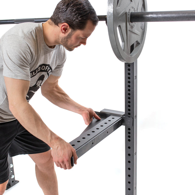 A man in a gray T-shirt and black shorts adjusts the height of the heavy-duty Bells of Steel weightlifting rack, featuring Spotter Arms Rack Attachment with UHMW lining. A barbell and weight plates are set against a plain white background.