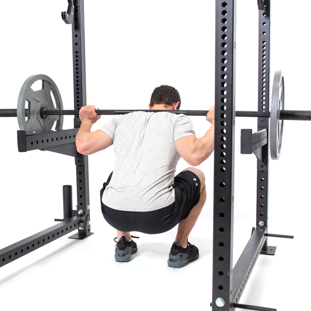 A person in a gray shirt and black shorts performs a squat with a barbell on their shoulders inside the Bells of Steel Spotter Arms Rack Attachment, featuring UHMW lining for extra protection.