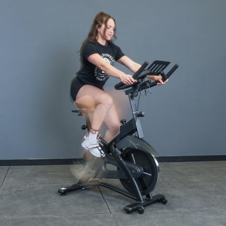 A woman in a black t-shirt and shorts rides the Bells of Steel Blitz Indoor Cycling Bike with magnetic resistance indoors against a plain gray wall, her legs blurred in motion.