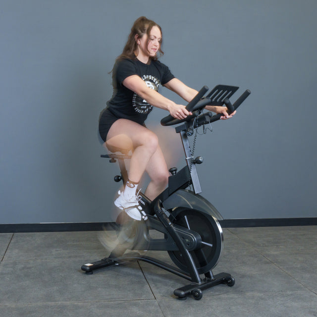 A woman in a black t-shirt and shorts rides the Bells of Steel Blitz Indoor Cycling Bike with magnetic resistance indoors against a plain gray wall, her legs blurred in motion.