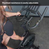 A person adjusts the magnetic resistance knob on the Bells of Steel Blitz Indoor Cycling Bike. The display screen and adjustable handlebars are visible. Text at the top reads, "Flywheel resistance is easily adjustable.