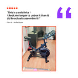 A Bells of Steel Blitz Indoor Cycling Bike with adjustable handlebars is on a wooden floor in a living room by a fireplace. A customer review above the image praises its easy assembly and sturdy build.