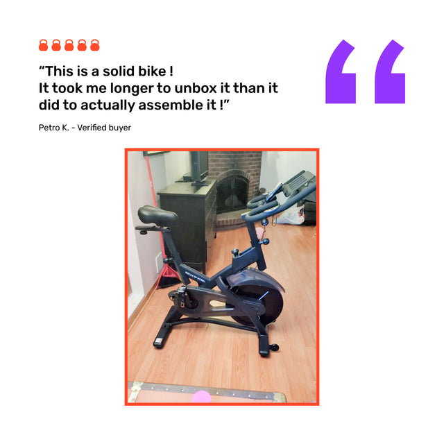 A Bells of Steel Blitz Indoor Cycling Bike with adjustable handlebars is on a wooden floor in a living room by a fireplace. A customer review above the image praises its easy assembly and sturdy build.
