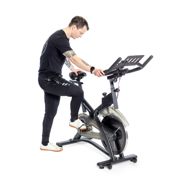 Dressed in black workout gear and white sneakers, a person adjusts the handlebars of a Blitz Indoor Cycling Bike by Bells of Steel, known for its magnetic resistance and sleek design, all set against a plain white background.