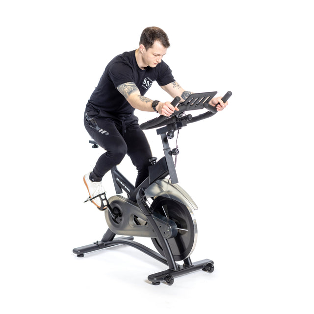 A person in a black t-shirt and pants is intensely riding the Bells of Steel Blitz Indoor Cycling Bike, gripping its adjustable handlebars with focus. The stationary bike, featuring magnetic resistance, is set against a plain white background in the side-view image.