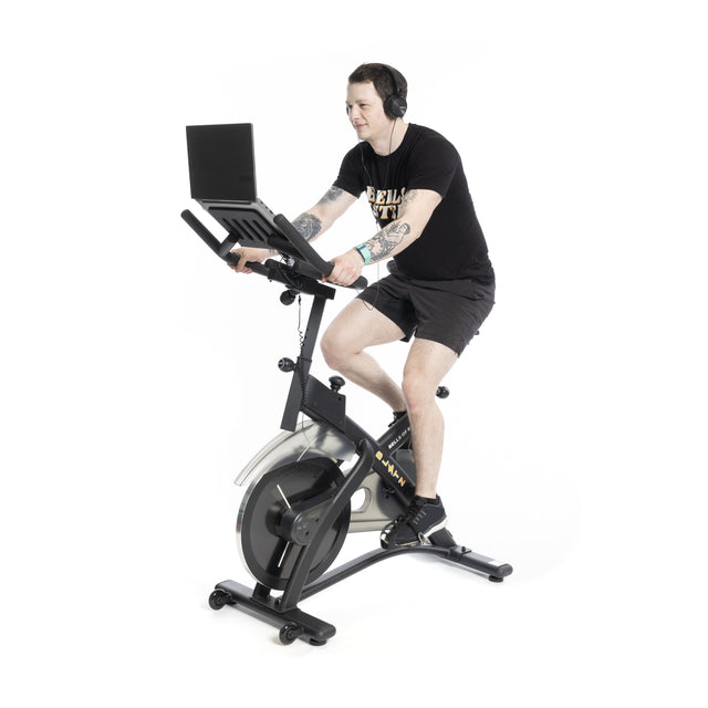 A person with tattoos wears headphones, a black t-shirt, and shorts while riding a Bells of Steel Blitz Indoor Cycling Bike with magnetic resistance and adjustable handlebars, using a laptop on the bike's stand against a plain white background.