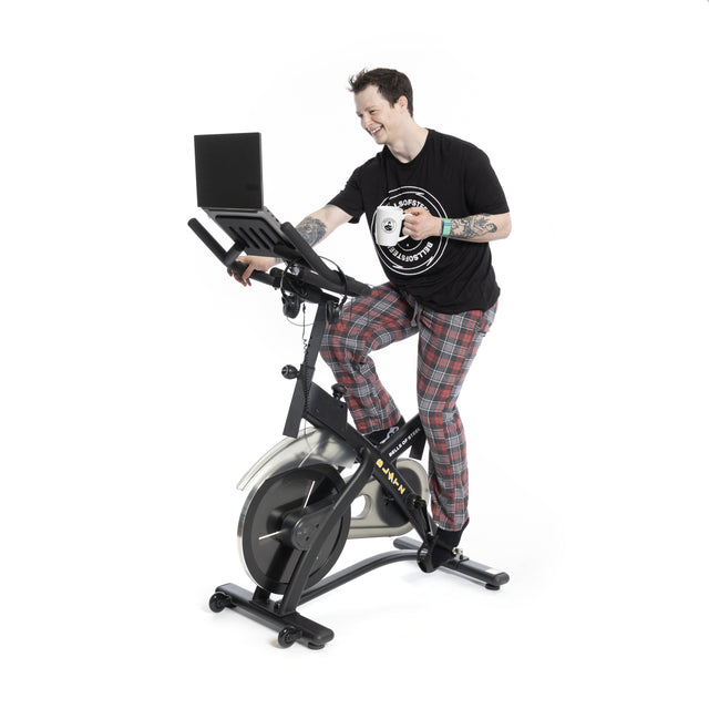 Wearing plaid pajama pants and a black t-shirt, the person on Bells of Steel's Blitz Indoor Cycling Bike, with adjustable handlebars, holds a coffee mug while smiling at their laptop on the bike's stand. The background is white.