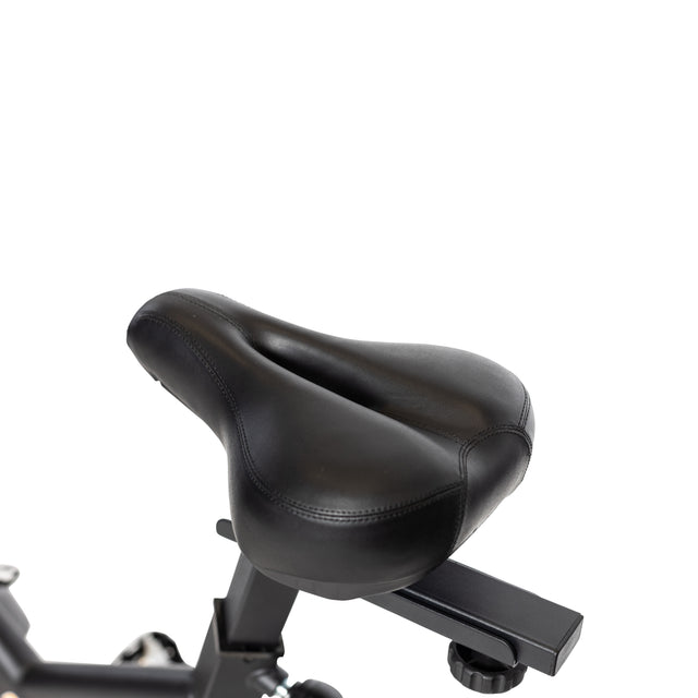 The Bells of Steel Blitz Indoor Cycling Bike features a close-up of its contoured black seat on a metal frame, isolated on a white background. The seat is designed for comfort and adjustable via an underneath knob.