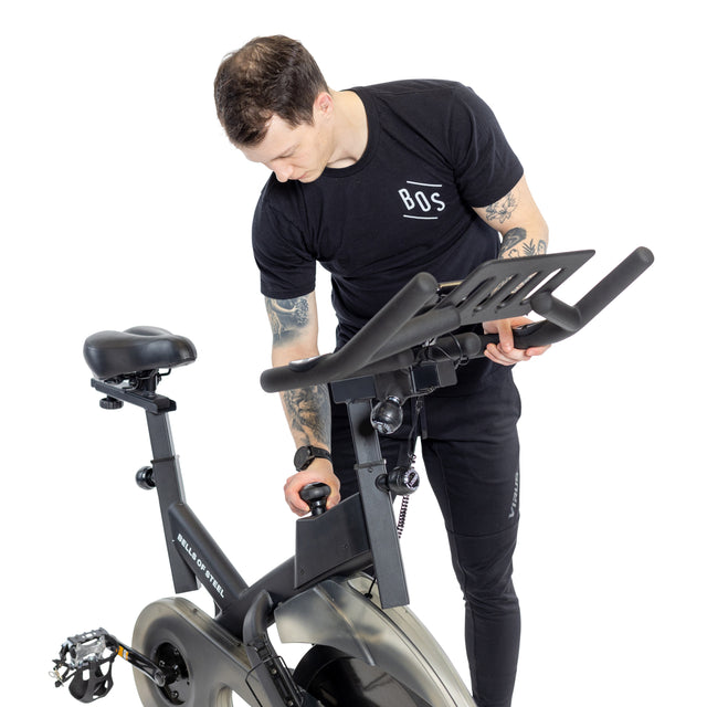 A person in a black t-shirt and dark pants adjusts the seat of a Bells of Steel Blitz Indoor Cycling Bike, which has adjustable handlebars and magnetic resistance. In a well-lit setting, the bike showcases its digital display and modern design.