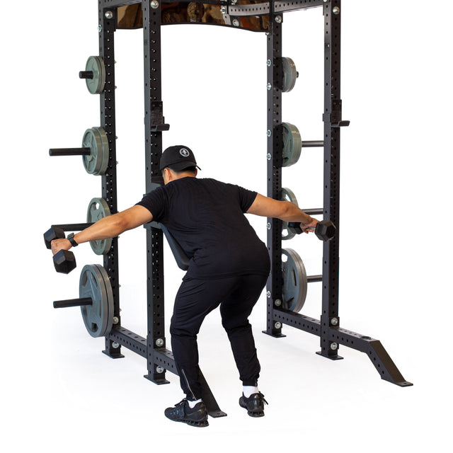 Wearing workout attire, a person performs a bent-over dumbbell lateral raise using the Seal Row Pad Rack Attachment by Bells of Steel for chest support. They hold a dumbbell in each hand, with their knees slightly bent, in front of a weight rack loaded with plates.