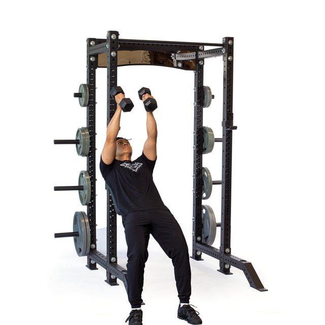 Dressed in black workout gear, a man executes a squat while gripping dumbbells. Positioned in front of him is a power rack equipped with weight plates stored on its side and features the Bells of Steel Seal Row Pad Rack Attachment as chest support. The background remains a plain white.