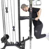 Wearing a black shirt, gray cap and pants, and white shoes, a person performs a tricep pushdown on the Bells of Steel All-in-One Trainer with various weights and pulleys on display.