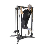 A person in a black shirt and cap is doing a pull-up on the Bells of Steel All-in-One Trainer, lifting with arms extended and legs bent. They wear sneakers and gray pants against a plain white background.