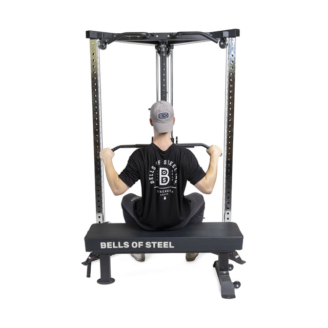 A person in a black Bells of Steel shirt and gray cap performs a seated lat pulldown on the Bells of Steel All-in-One Trainer, featuring adjustable height settings, a padded bench, and a squat stand for functional training.