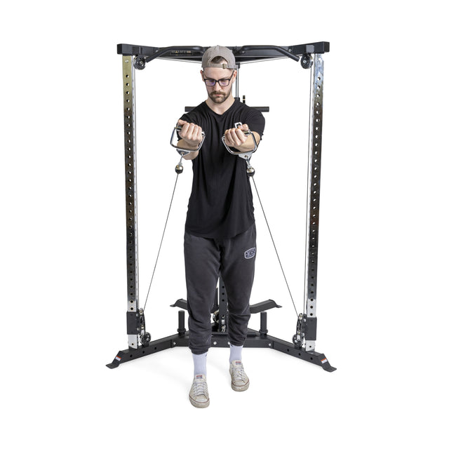 A man in glasses and a cap expertly uses the Bells of Steel All-in-One Trainer's cable crossover feature. He's dressed in a black t-shirt and gray pants, working out against a plain white background.