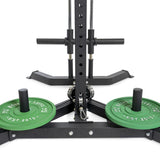 A close-up of the All-in-One Trainer features green 10 kg weight plates labeled "Bells of Steel." The image highlights the sturdy base and squat stand-like support, emphasizing its durability and reliability in strength training.