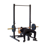 Wearing black, a person uses the Hydra Squat Stand Builder by Bells of Steel, a sturdy black rack with modular capability, for their bench press exercise. They lie on a bench under this versatile squat stand while lifting a barbell with weights on both sides in their home gym.