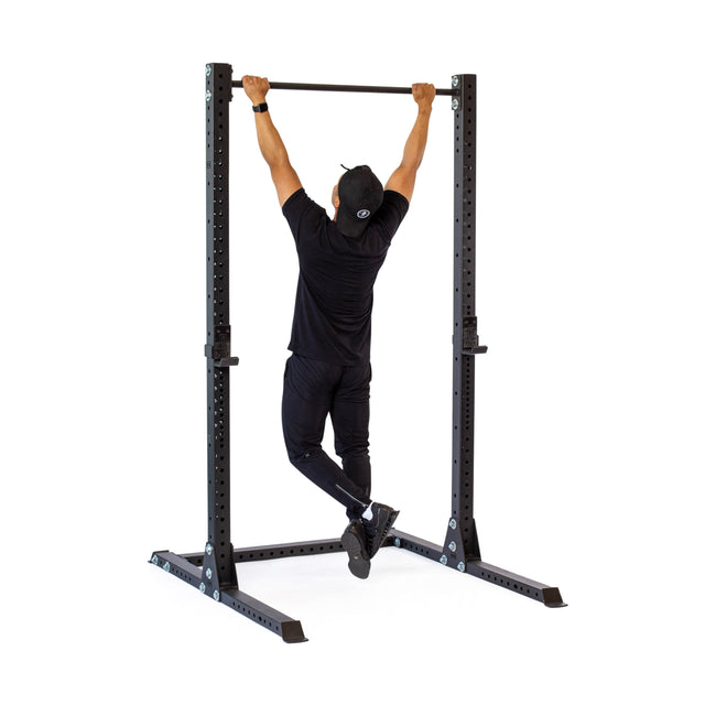 A person in black athletic wear and a cap performs a pull-up on the freestanding Bells of Steel Hydra Squat Stand - Prebuilt, ideal for home gyms. The plain white background accentuates the exercise equipment and their focused position.