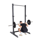 Poised in a power rack wearing a black shirt, cap, and shorts, a person utilizes the Bells of Steel Manticore Squat Stand Builder (3" x 3", 1" Holes) under its impressive 1,000-pound capacity against a plain white background.