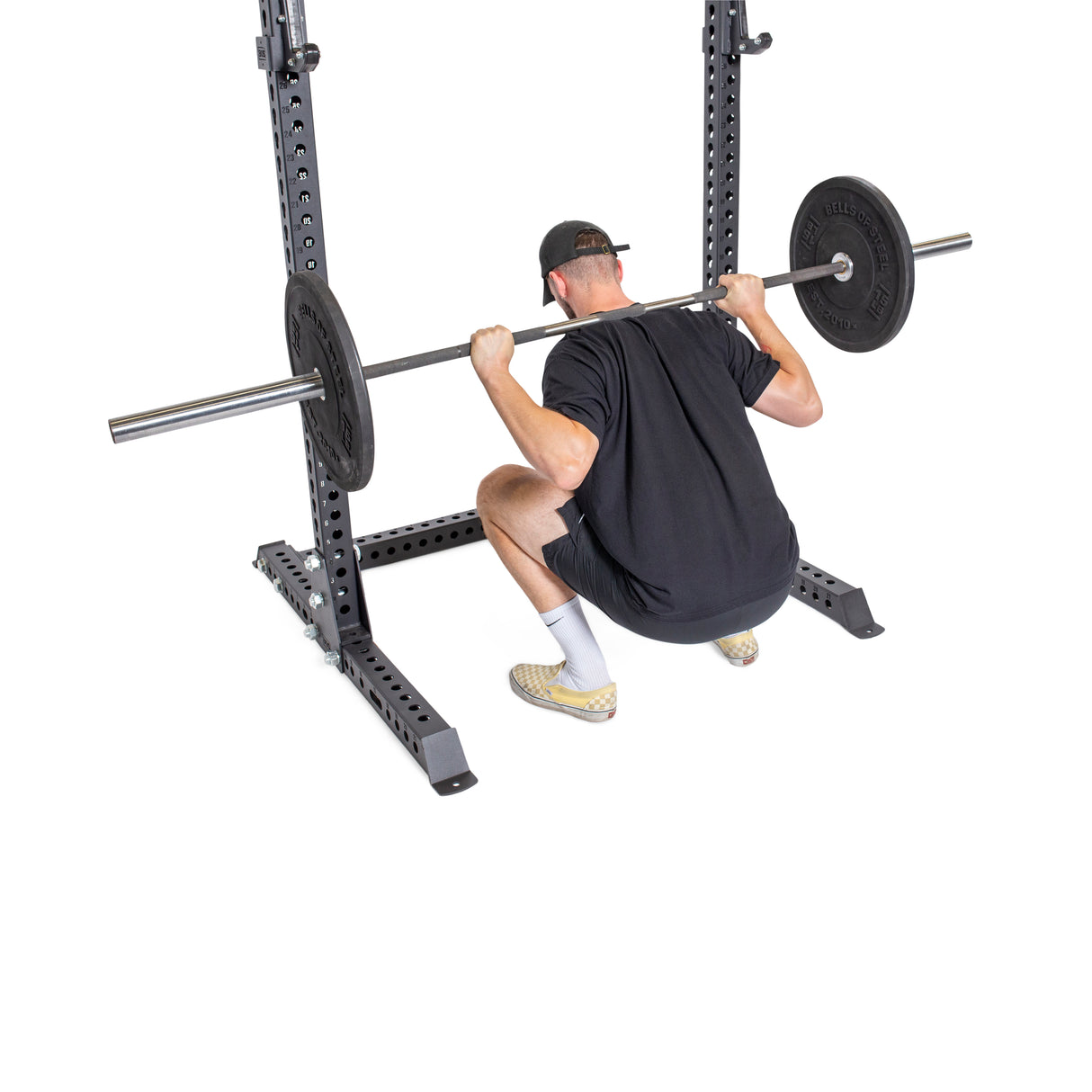 A person in sportswear squats with a barbell on a Bells of Steel Manticore Squat Stand Builder, which has a 1,000-pound capacity. They're in a black shirt, black cap, and light shoes against a plain white background.