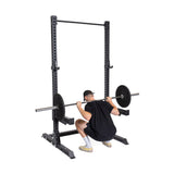 A person squats with a barbell in the Bells of Steel Manticore Squat Stand Builder, featuring 3" x 3" adjustable holes. They wear a black shirt, shorts, and cap. The barbell's weights are securely fastened.