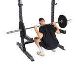 A man in a black shirt and shorts performs a squat with a weighted barbell on his shoulders, using the Bells of Steel Manticore Squat Stand Builder (3" x 3", 1" Holes). He wears a cap and athletic shoes, all set against a white background.