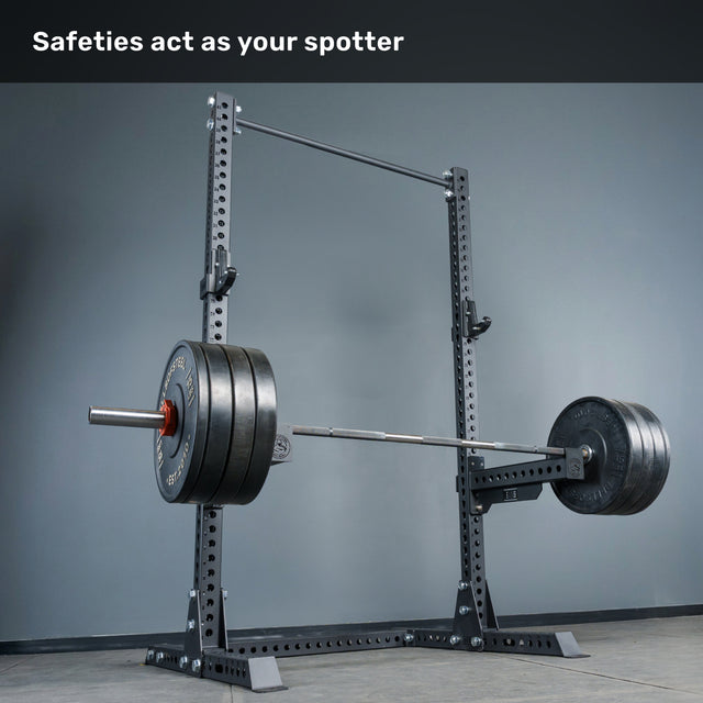 A Bells of Steel Manticore Squat Stand - Prebuilt (3" x 3", 1" Holes), equipped with safety arms and a heavily loaded barbell, stands ready in a gym with gray walls and a concrete floor. The caption above reads, "Safeties act as your spotter." This robust squat stand, featuring durable 3x3 uprights, makes an excellent addition to any home gym setup.