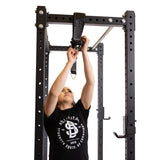 A person in a black t-shirt is reaching up to adjust the Bells of Steel Suspension Spotter Straps and Bodyweight System on a squat rack inside a gym.