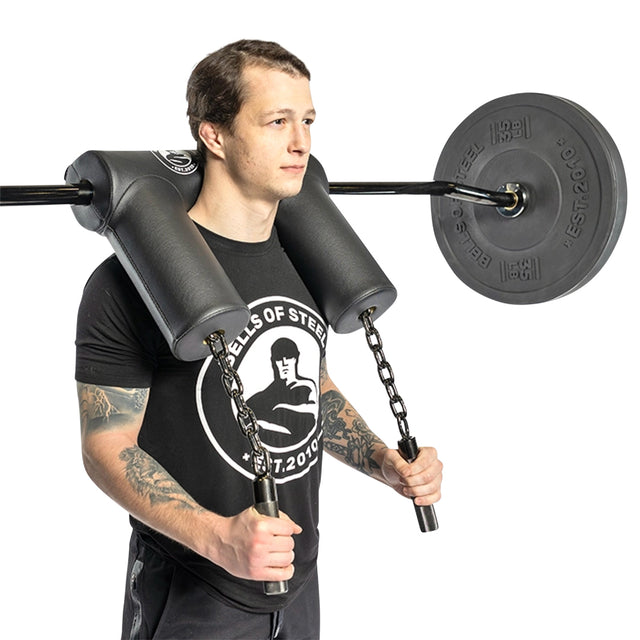 A person confidently stands with the Bells of Steel Safety Squat Bar - The SS4, wearing a black T-shirt and shorts. The ergonomic pad enhances comfort as they grip chains for versatile squat exercises.