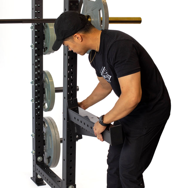 A person in a black outfit and cap adjusts the Bells of Steel Spotter Arms Rack Attachment on a loaded squat rack with UHMW lining for protection.