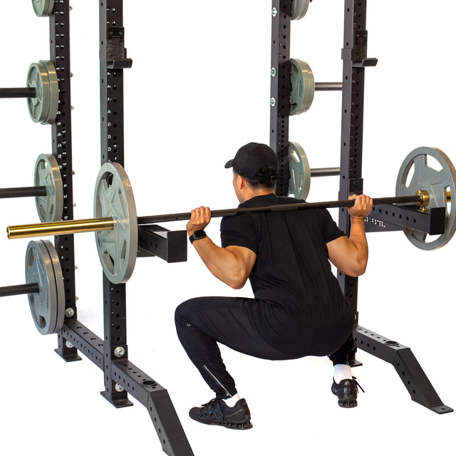 A person in black athletic wear and a cap performs a squat with a barbell on their shoulders, using the Bells of Steel Spotter Arms Rack Attachment. The squat rack, set against a plain white background, holds several weight plates.