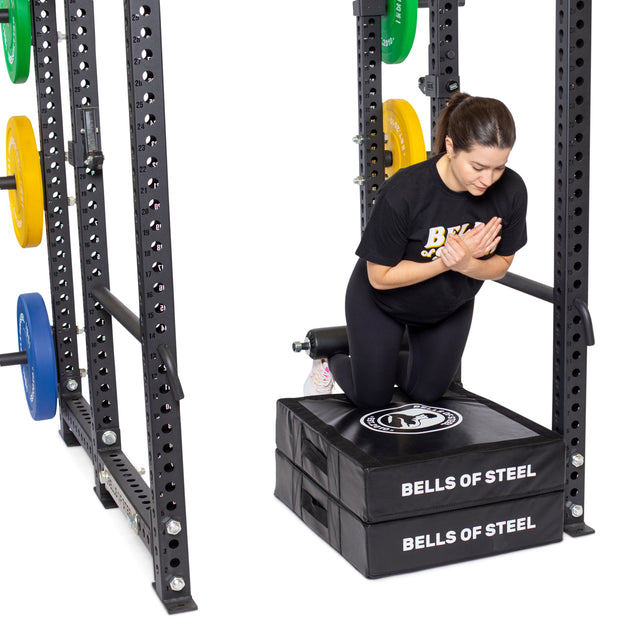 Kneeling on a padded platform inside a squat rack, a person performs Nordic hamstring curls with precision. The setup is enhanced by colorful weight plates and the Split Squat Leg Roller Rack Attachment. They're sporting a black "Bells of Steel" t-shirt.