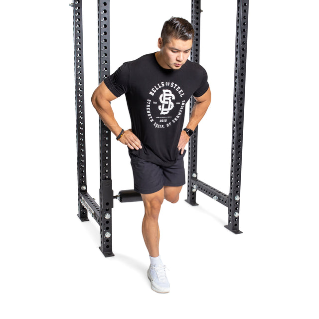 A person using the Bells of Steel Split Squat Leg Roller Rack Attachment performs a Bulgarian split squat with one leg forward, wearing a black T-shirt, shorts, and white athletic shoes.