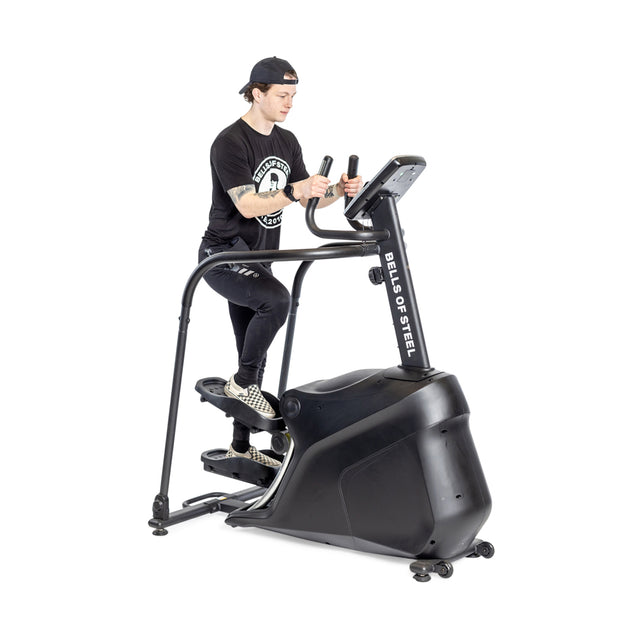 An individual dressed in a black cap, t-shirt, and leggings is working out on a Bells of Steel Blitz Stepper elliptical machine. Utilizing its magnetic resistance for joint-friendly cardio, they grip the handles tightly and concentrate on the console screen.