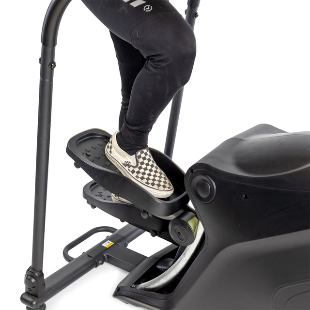 A person wearing black pants and checkered slip-on sneakers steps onto the Bells of Steel Blitz Stepper, highlighting their legs and feet as one foot rests on the joint-friendly cardio machine's surface. This equipment offers smooth magnetic resistance for an efficient workout.